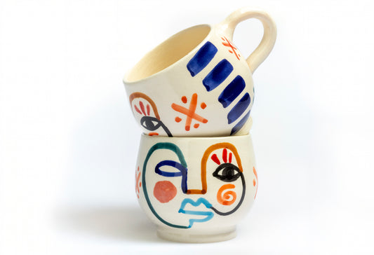 Two ceramic mugs with cream-colored bodies featuring hand-painted abstract faces in blue, orange, and black geometric designs