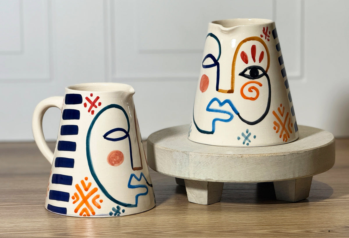 Ceramic pitcher and mug with hand-painted abstract faces in blue, orange, and red on cream background.