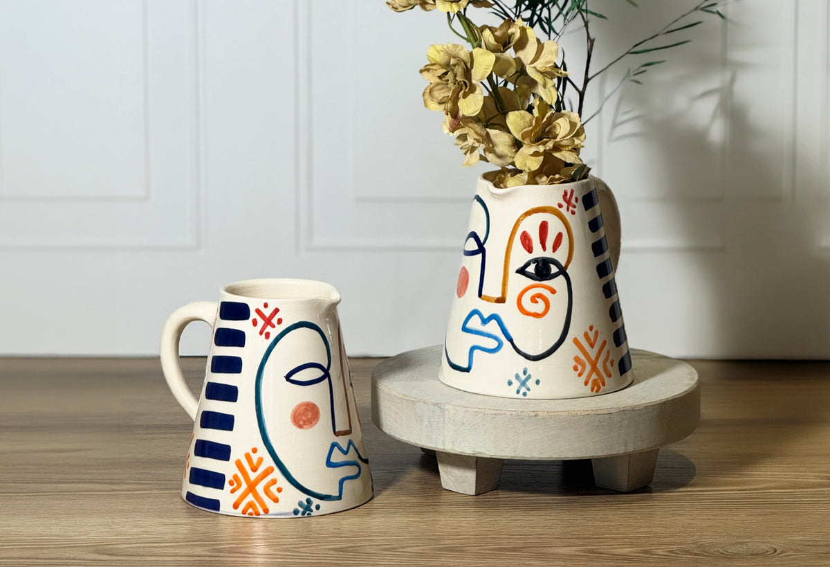 Cream ceramic pitcher with hand-painted abstract face design in navy, orange, and red, holding dried flowers.