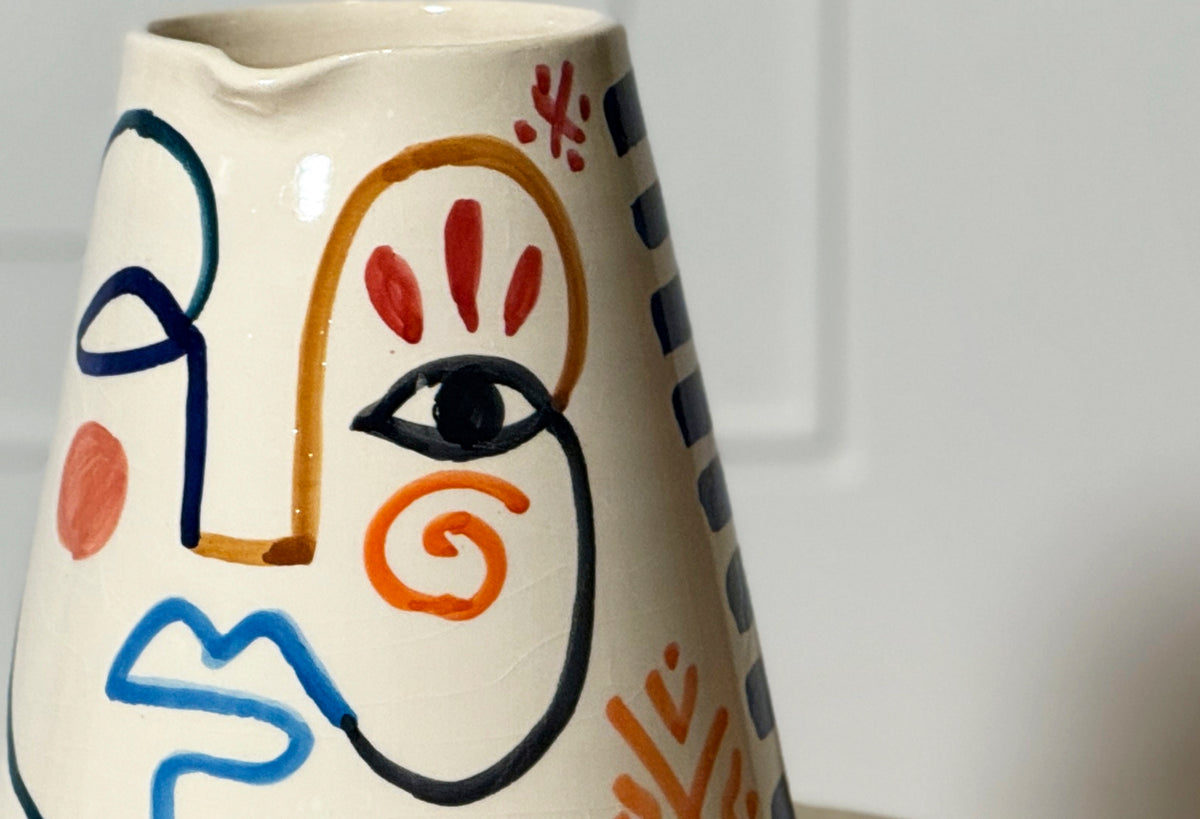 Cream ceramic pitcher with hand-painted abstract face featuring blue, orange, red, and black lines and shapes.