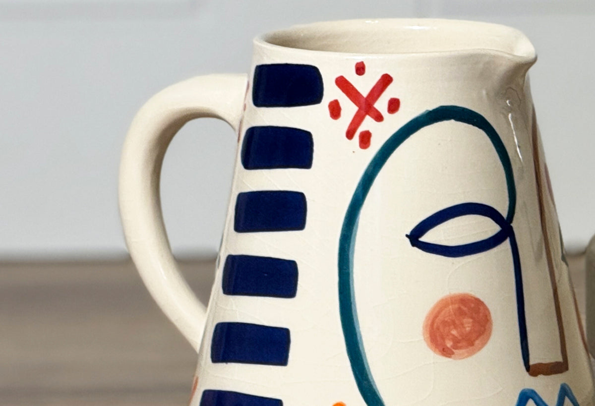 Cream ceramic pitcher with hand-painted abstract face featuring navy rectangles, red X mark, teal outline, and coral dot deta