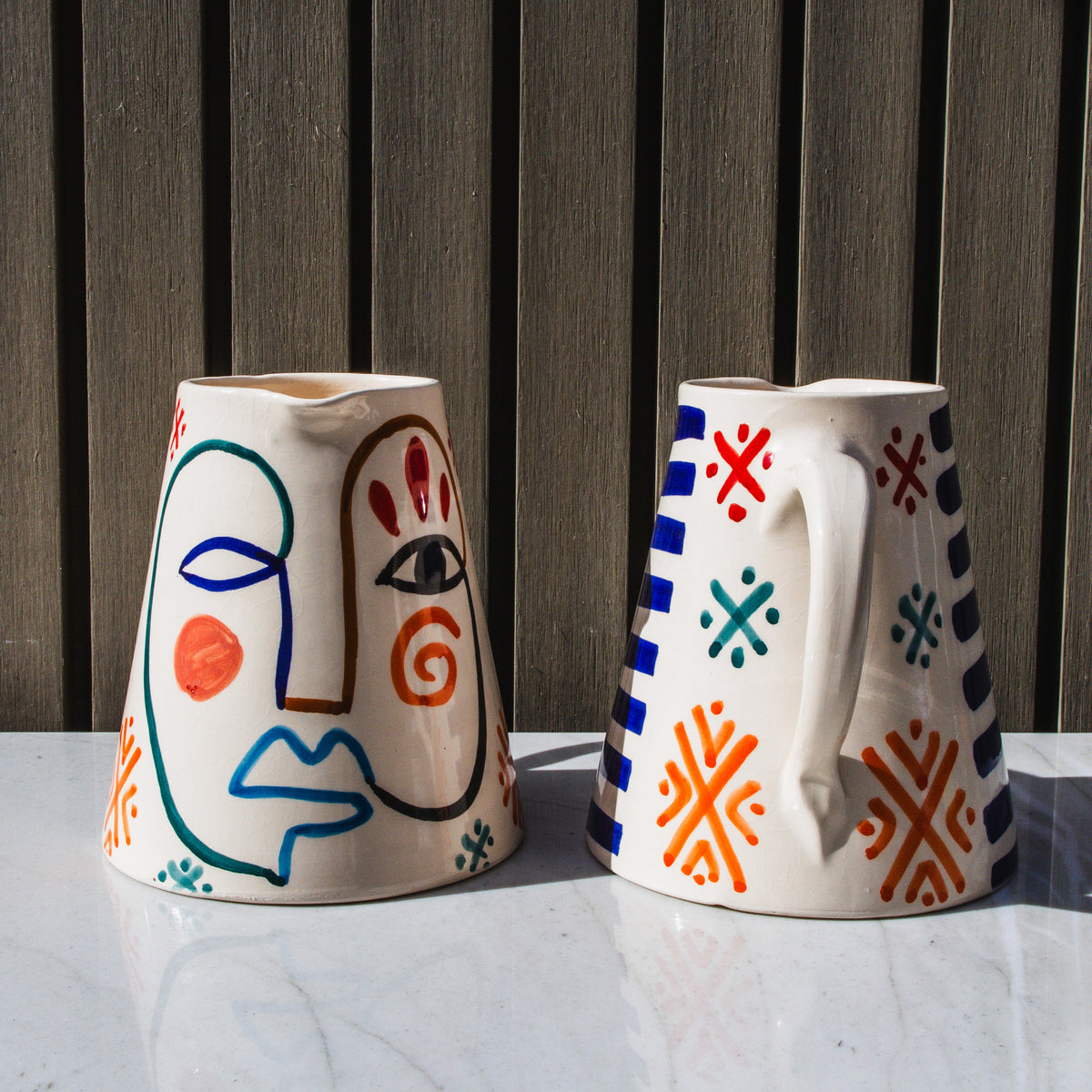 Two cream ceramic pitchers with hand-painted abstract faces and geometric patterns in blue, red, orange, and teal.