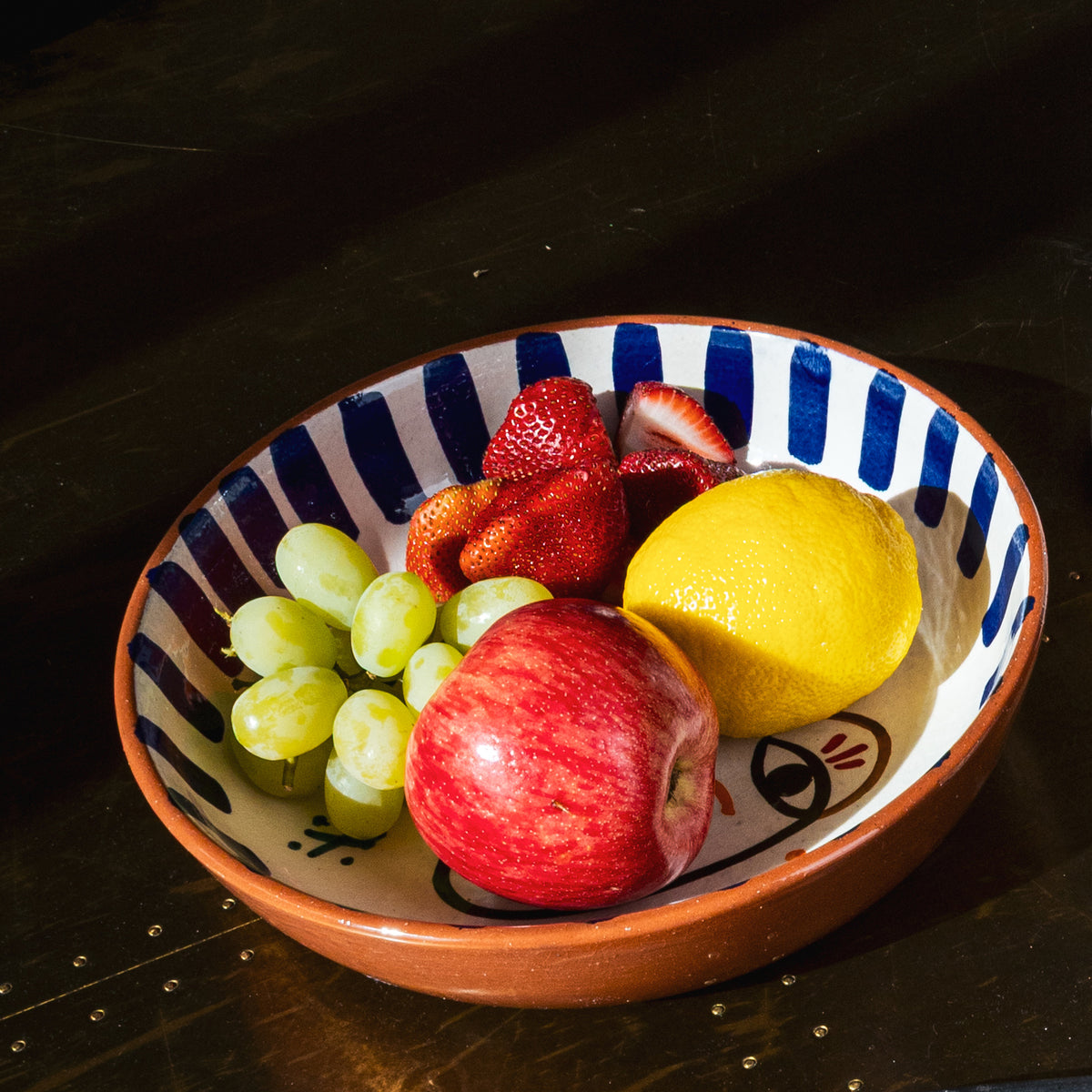 Ceramic bowl with blue and white striped interior, terracotta rim, and hand-painted facial features, filled with strawberries