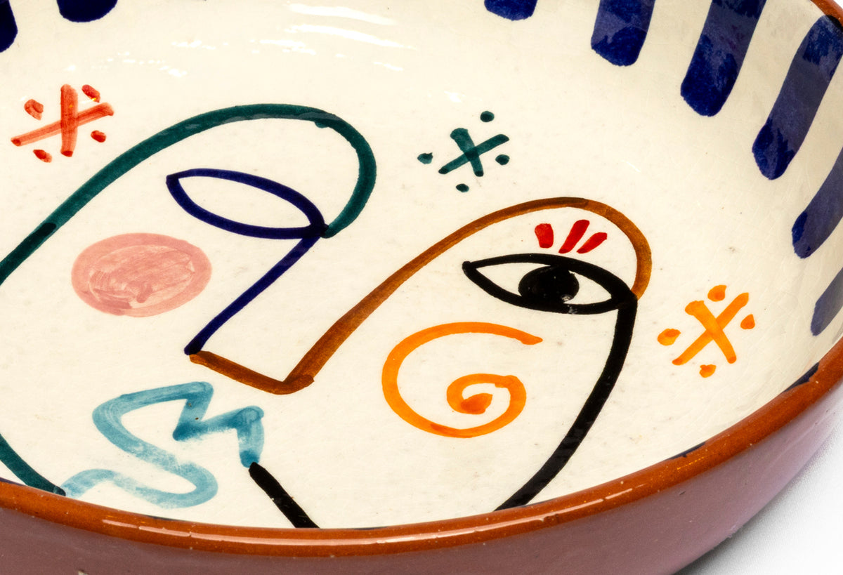 Ceramic bowl with hand-painted abstract face design in navy, orange, teal, and red on cream background.