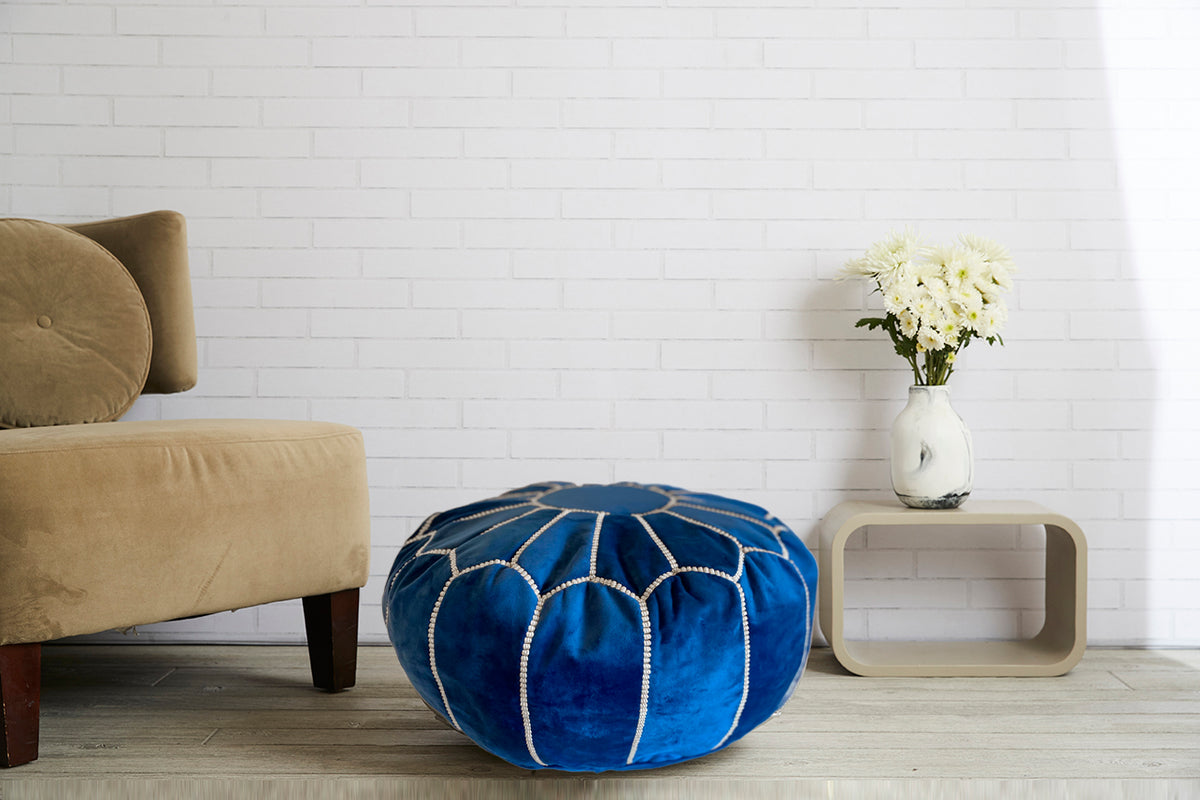 Blue velvet round pouf with white geometric stitching pattern on wooden floor next to chair.