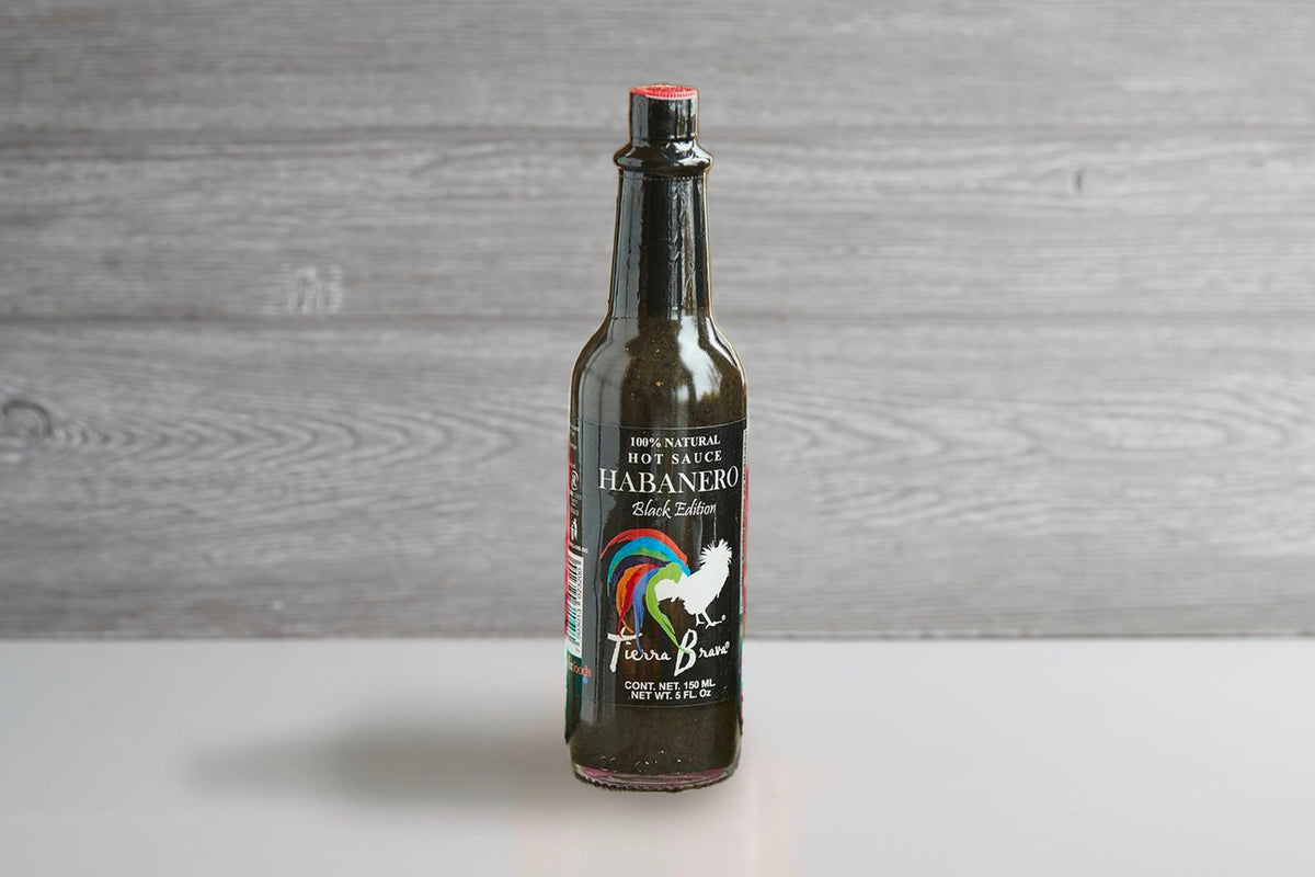 Dark glass bottle with red cap labeled "Tierra Brava Habanero" hot sauce on gray wooden background.