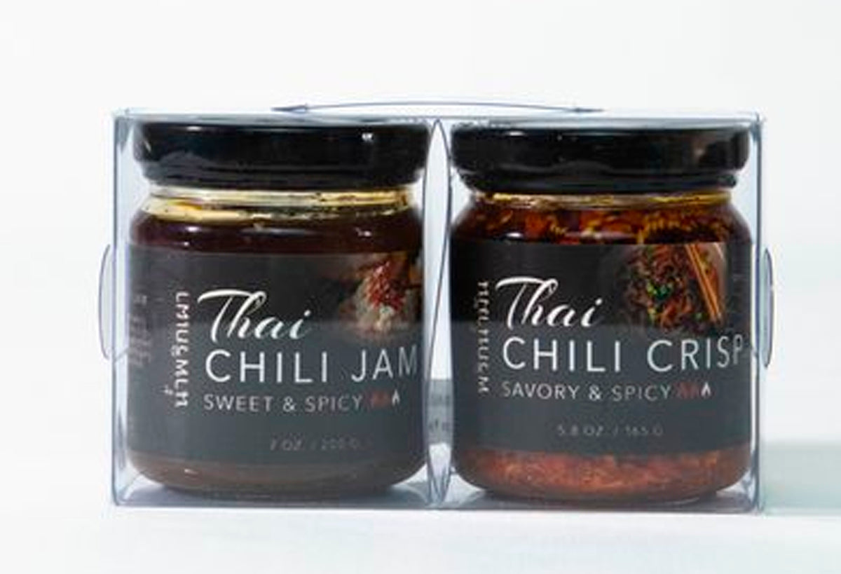 Two glass jars with black lids containing Thai chili jam and chili crisp in a clear plastic gift box.