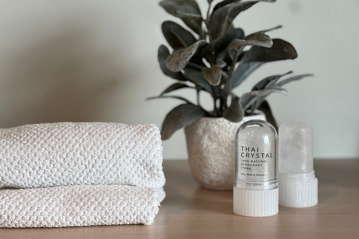 Two white Thai crystal deodorant stones with applicator caps displayed next to folded towels and potted plant.
