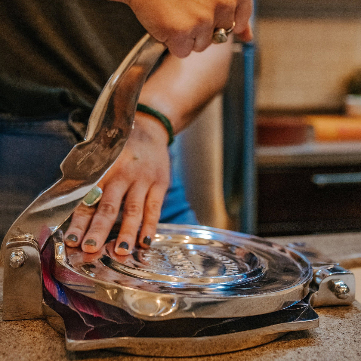 Polished aluminum tortilla press with lever handle being operated by a person's hands over a countertop.