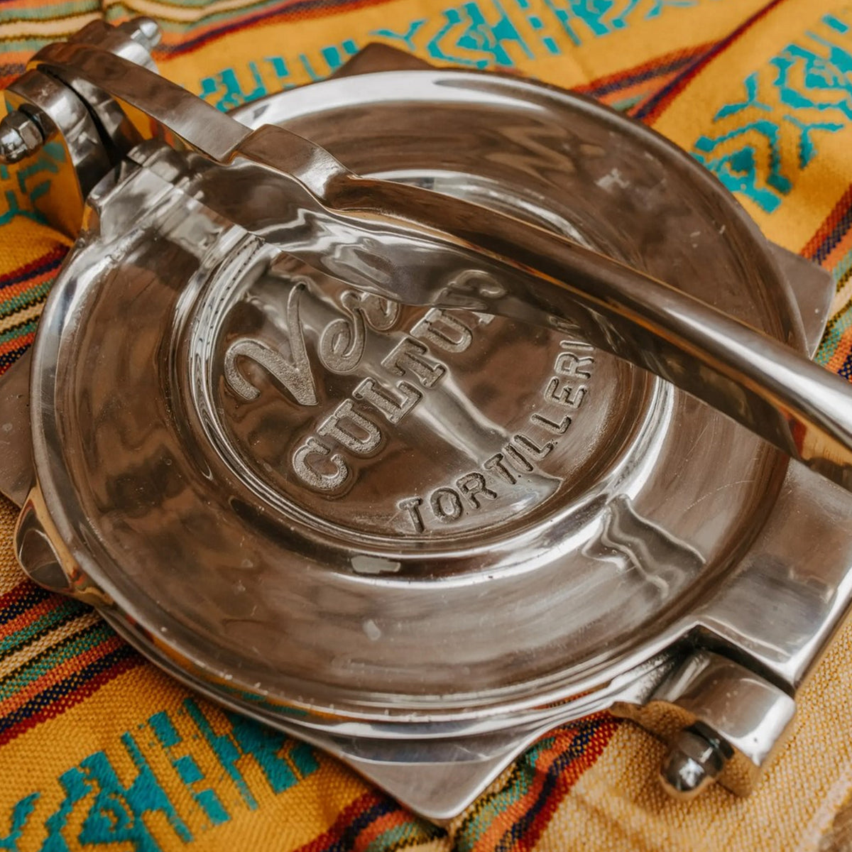 Polished aluminum tortilla press with lever handle and embossed text on circular pressing surface, shown open on colorful wov