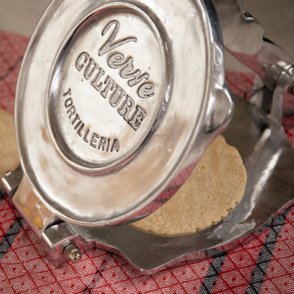 Polished aluminum tortilla press with "2 Ere Tortilleria" engraved on circular pressing plate, containing a flattened tortill