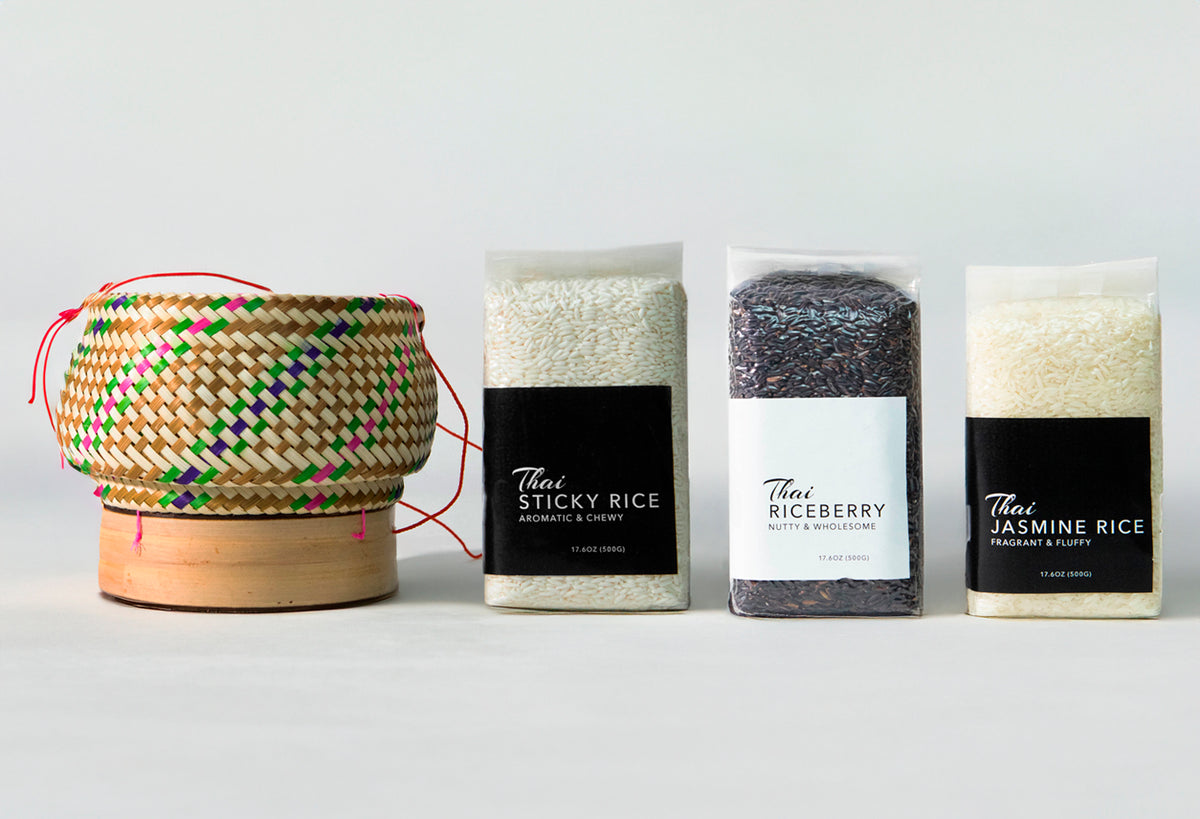 Woven rice basket with red handles next to three packaged rice varieties: sticky rice, riceberry, and jasmine rice.