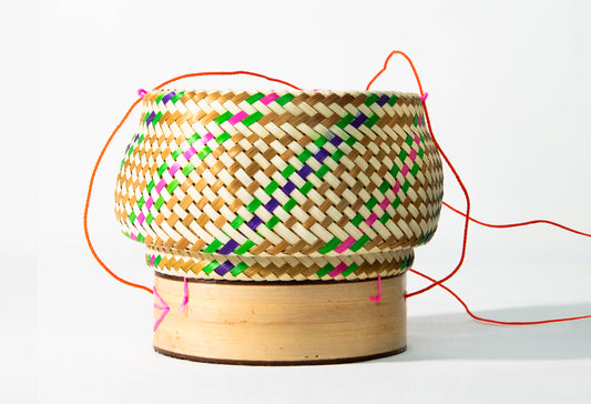 Woven bamboo basket with geometric pattern in green, pink, yellow and navy, wooden base, and orange fabric handles.
