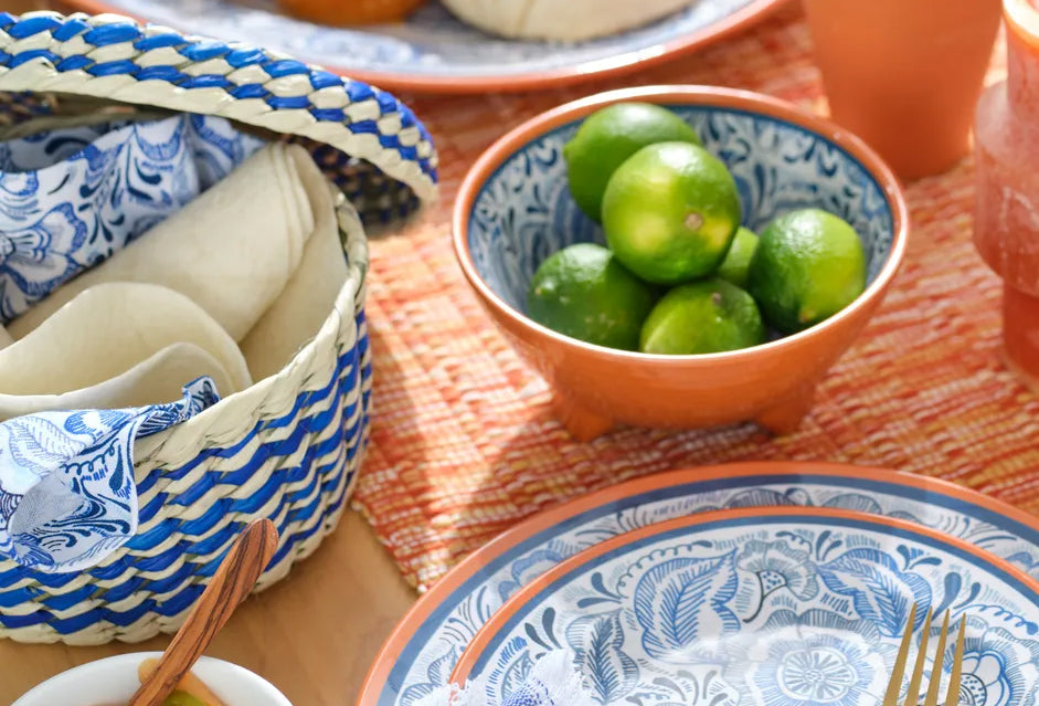 Woven palm basket with blue and cream wavy pattern containing folded tortillas, beside ceramic bowls and dinnerware.