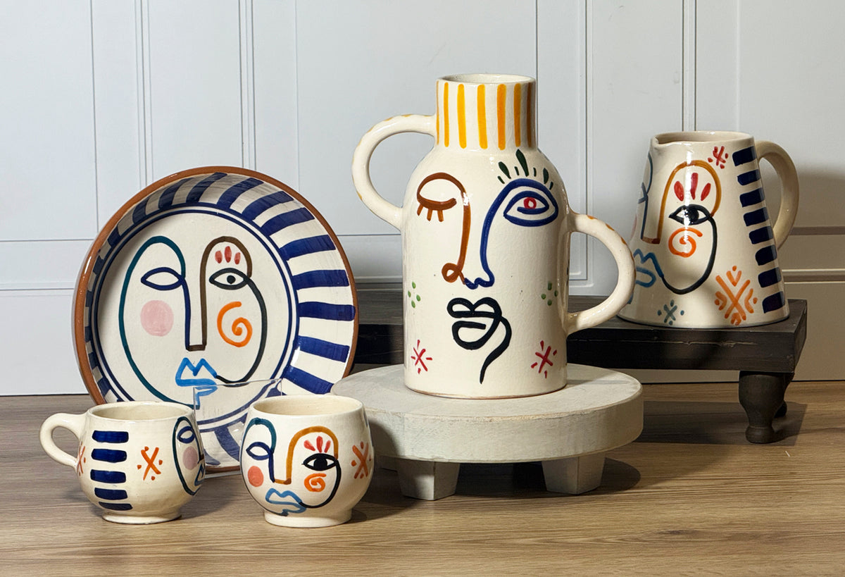 Two cream ceramic mugs with hand-painted abstract faces in blue, red, and orange lines and geometric patterns.