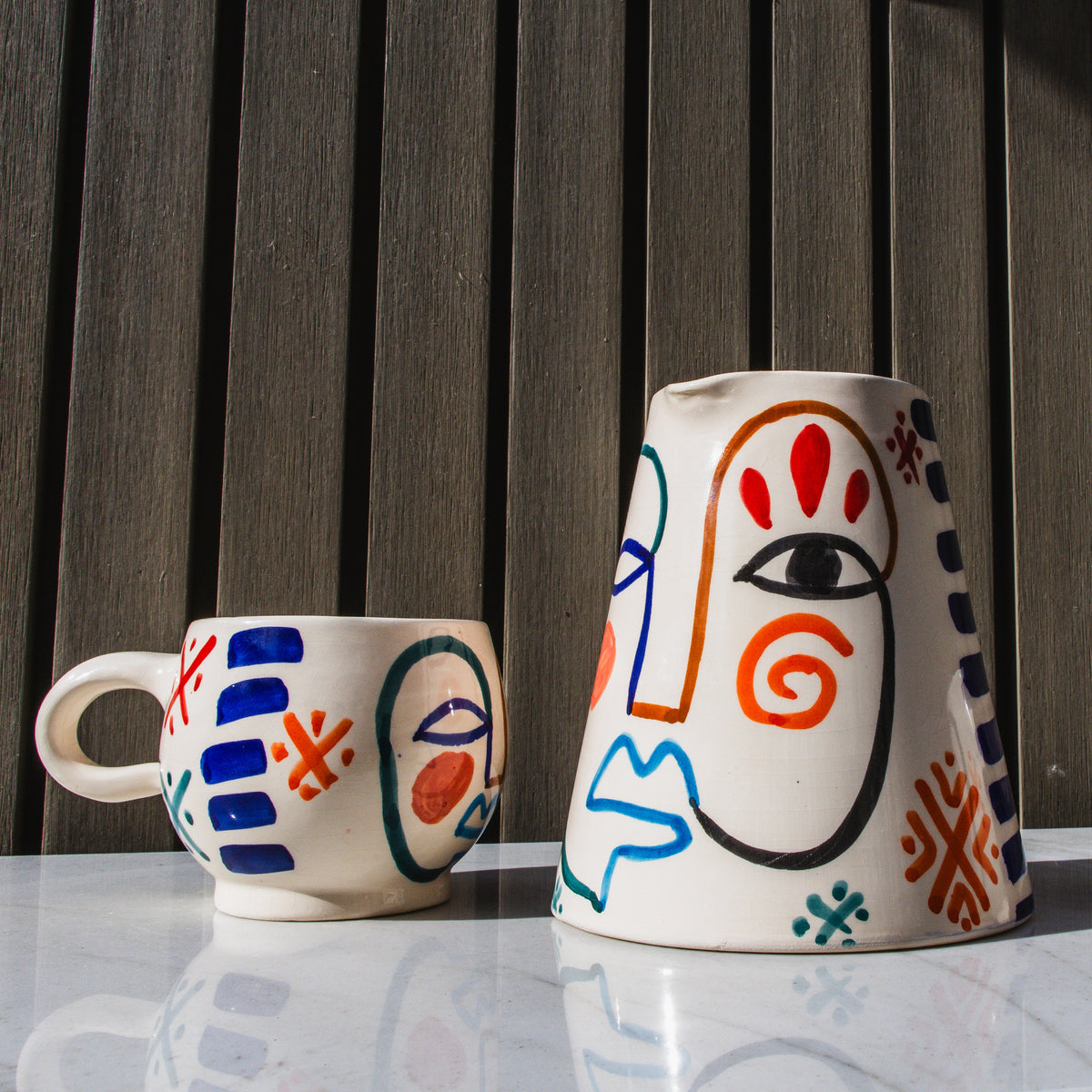 Cream ceramic pitcher with abstract face design in red, orange, blue, and black hand-painted details, shown next to matching 