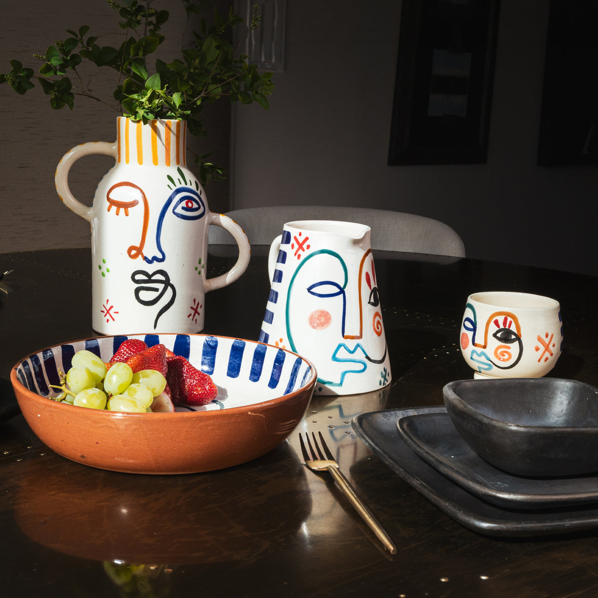 Cream ceramic pitcher with hand-painted abstract faces in blue, orange, and black with striped rim and two handles.