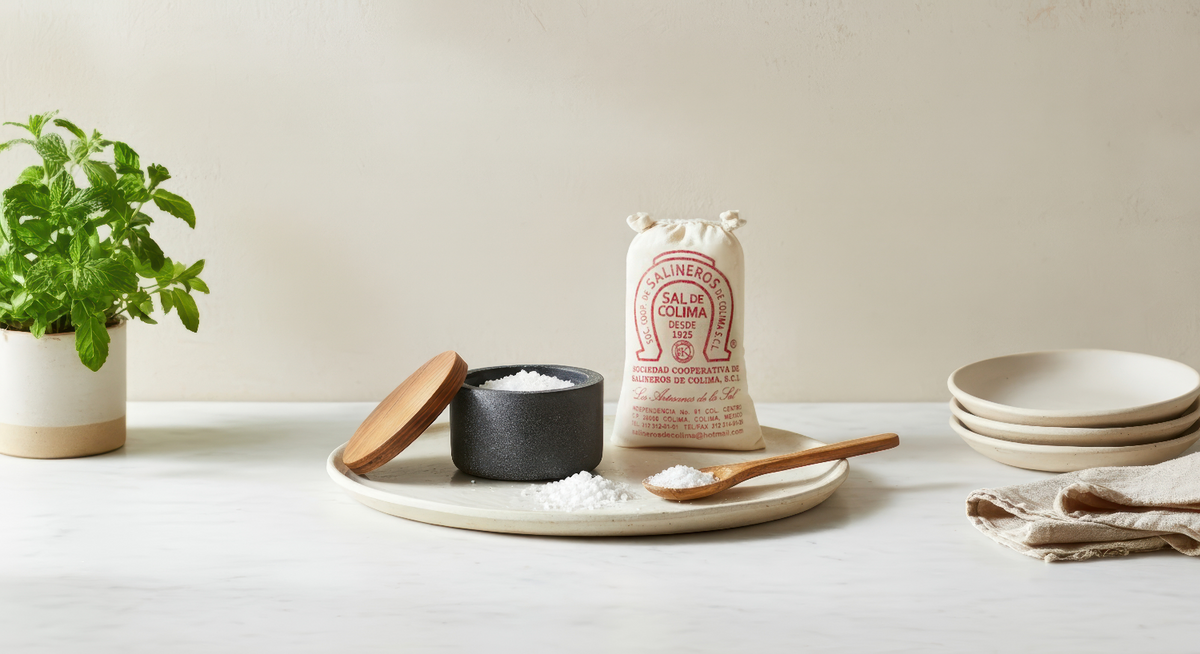 White sea salt in black ceramic bowl with wooden spoon on beige plate, burlap bag in background, fresh herbs and bowls nearby