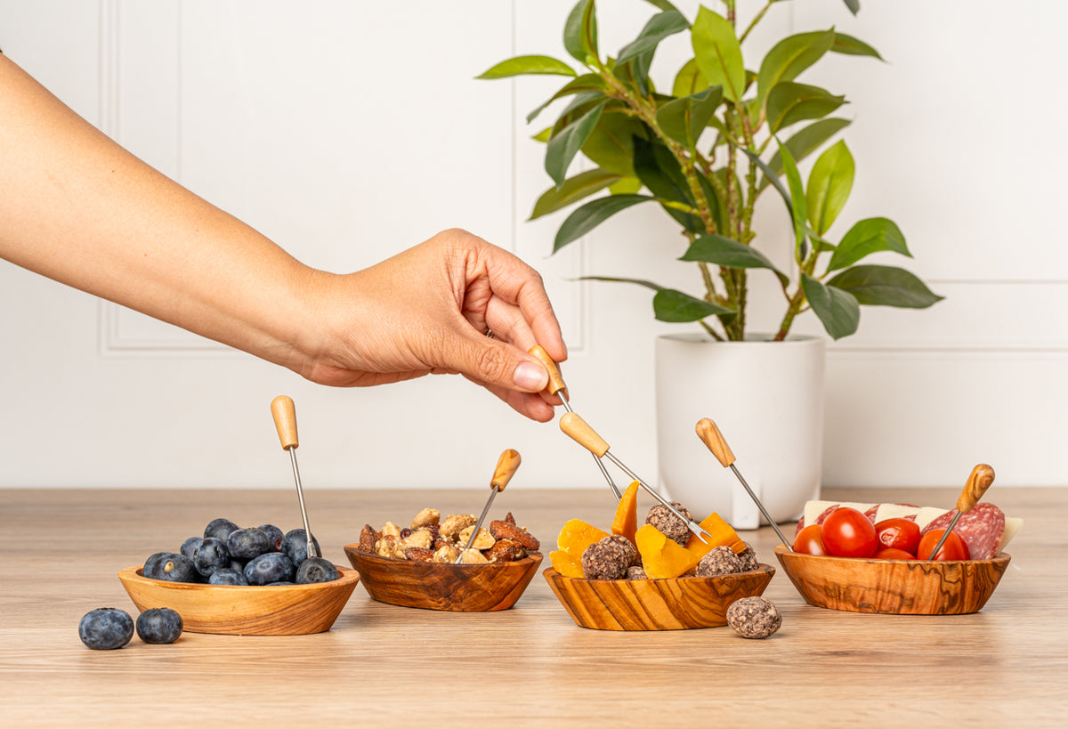 Six small forks with olive wood handles and metal tines displayed in wooden serving bowls with appetizers.