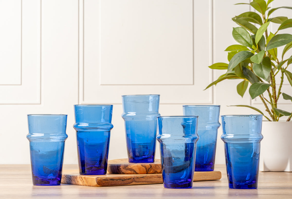 Six blue Moroccan Beldi glasses with textured surfaces arranged on wooden surface with potted plant.
