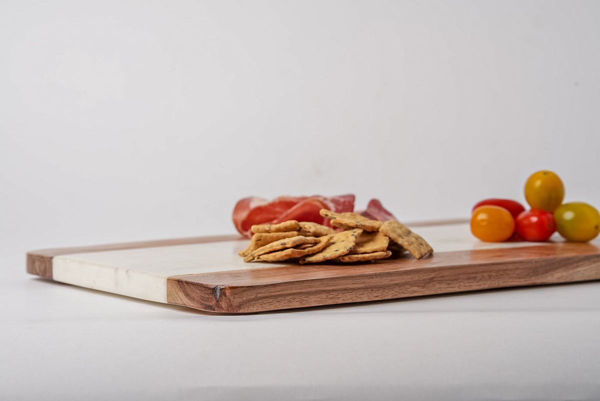 Rectangular serving board with white marble section and dark wood border, topped with crackers, cured meat, and colorful grap