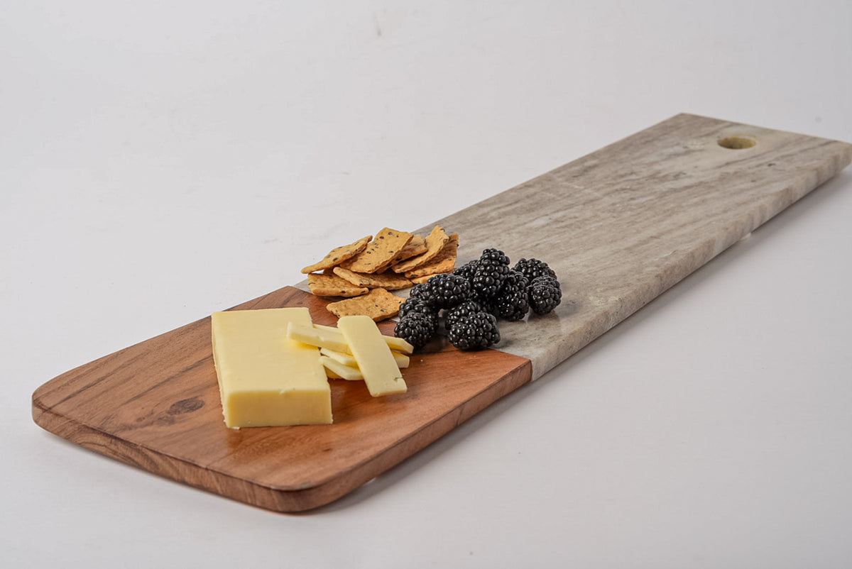 Long rectangular charcuterie board with tan marble top section and acacia wood base, topped with cheese, crackers, and berrie