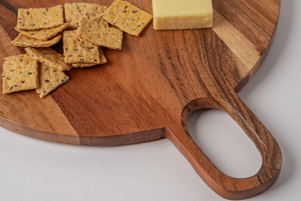 Round acacia wood serving board with handle cutout, displaying crackers and cheese.