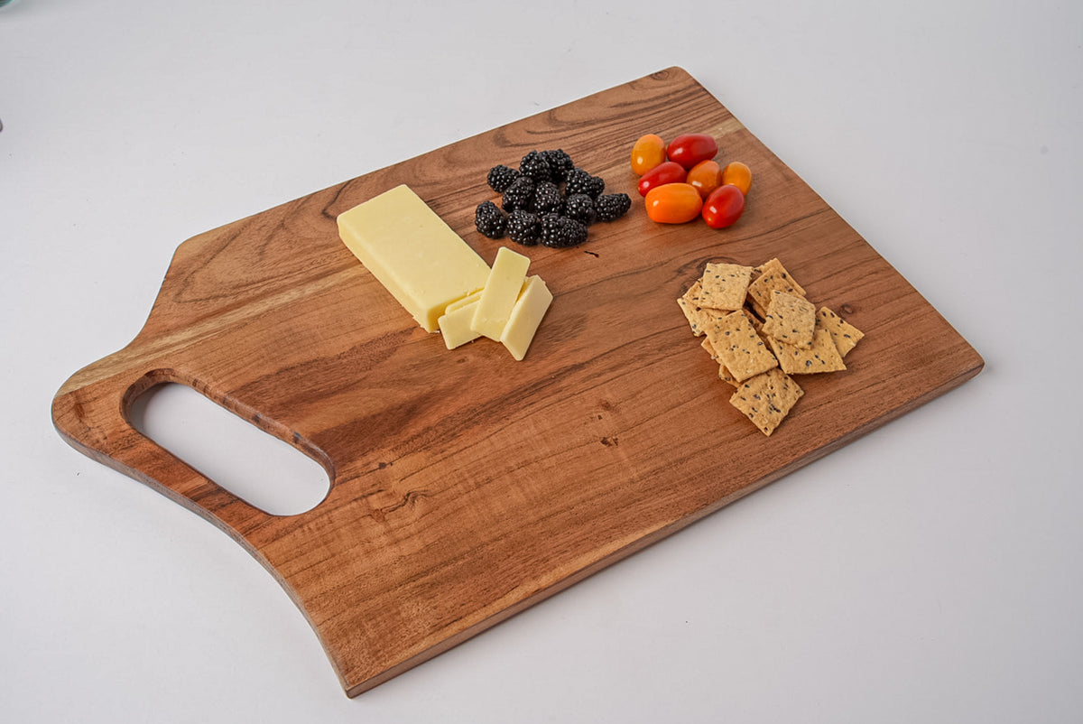 Brown acacia wood rectangular serving board with handle, displaying cheese, crackers, berries, and cherry tomatoes.