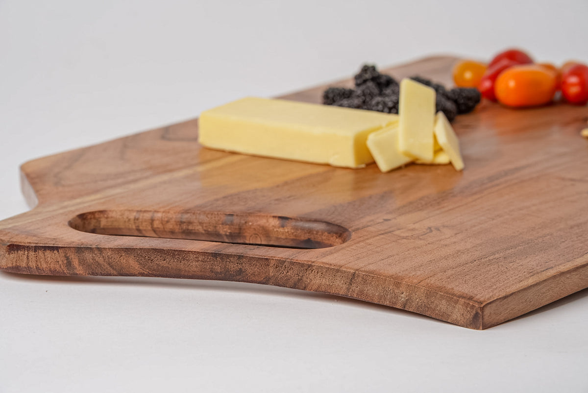 Rectangular acacia wood serving board with handle cutout, topped with cheese slices, berries, and tomatoes.