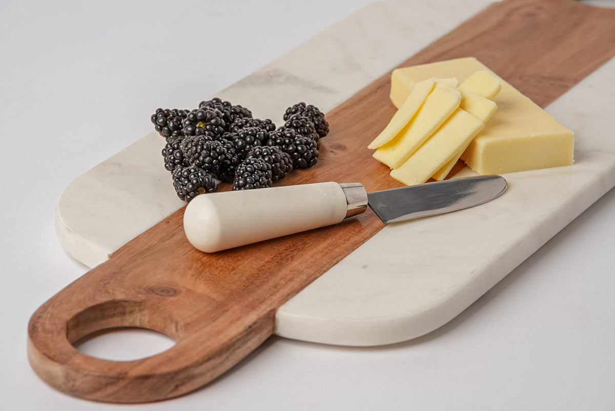 Rectangular marble and wood serving board with hole handle, displaying blackberries, cheese slices, and butter knife.