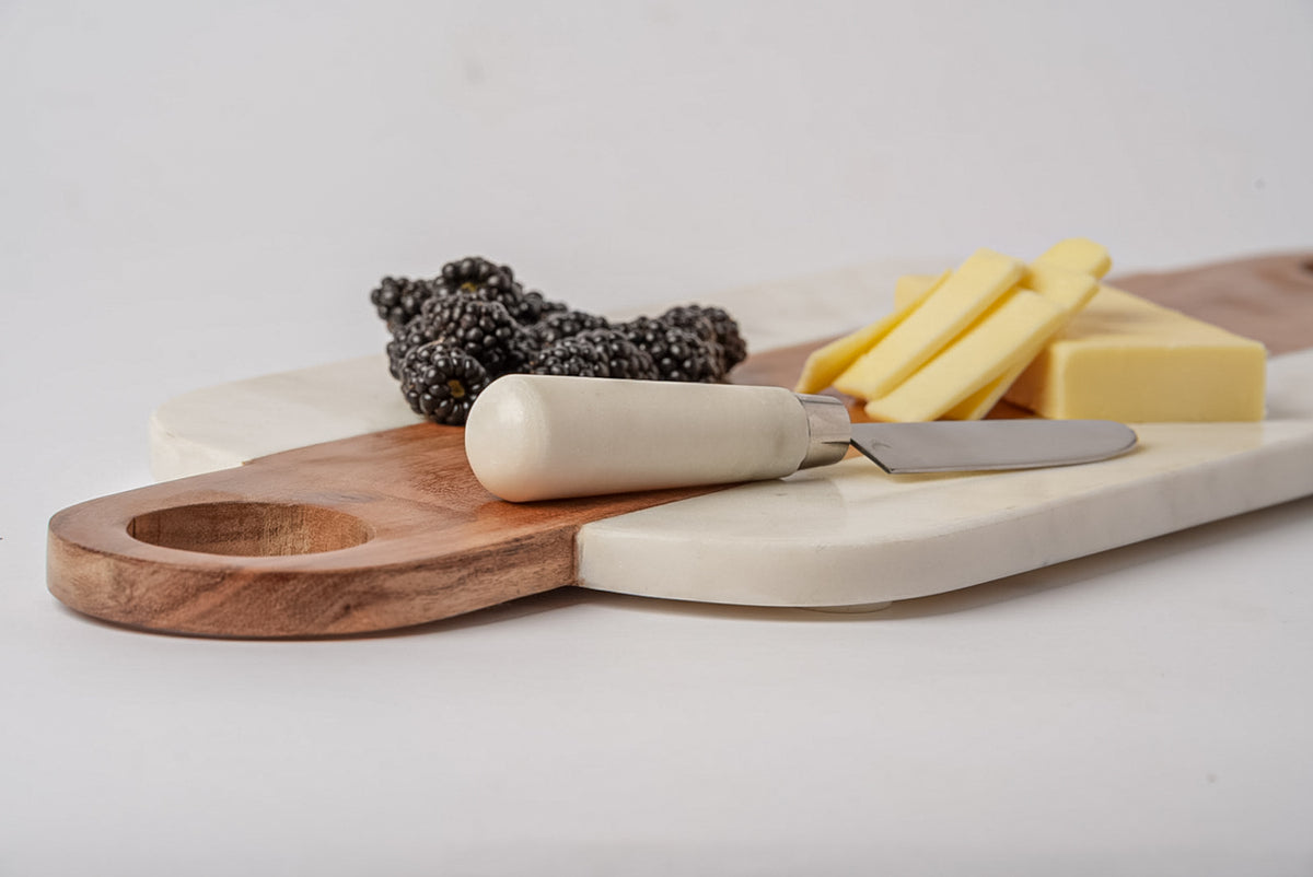 White marble serving board with wood handle section holding cheese, cured meats, and spreader knife.