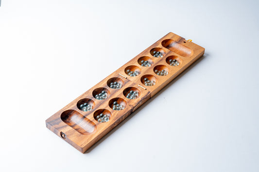 Wooden Mancala board with two end cups and multiple circular playing pits filled with small stones.