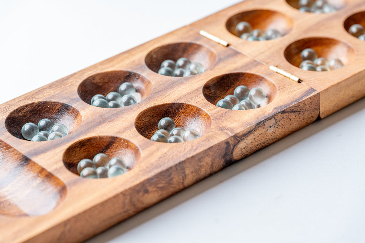 Wooden mancala board with multiple cup-shaped indentations filled with metal game pieces.