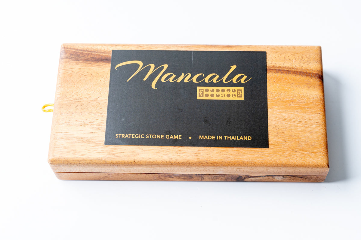 Wooden mancala game board with black label, multiple cup indentations, and storage compartment on side.