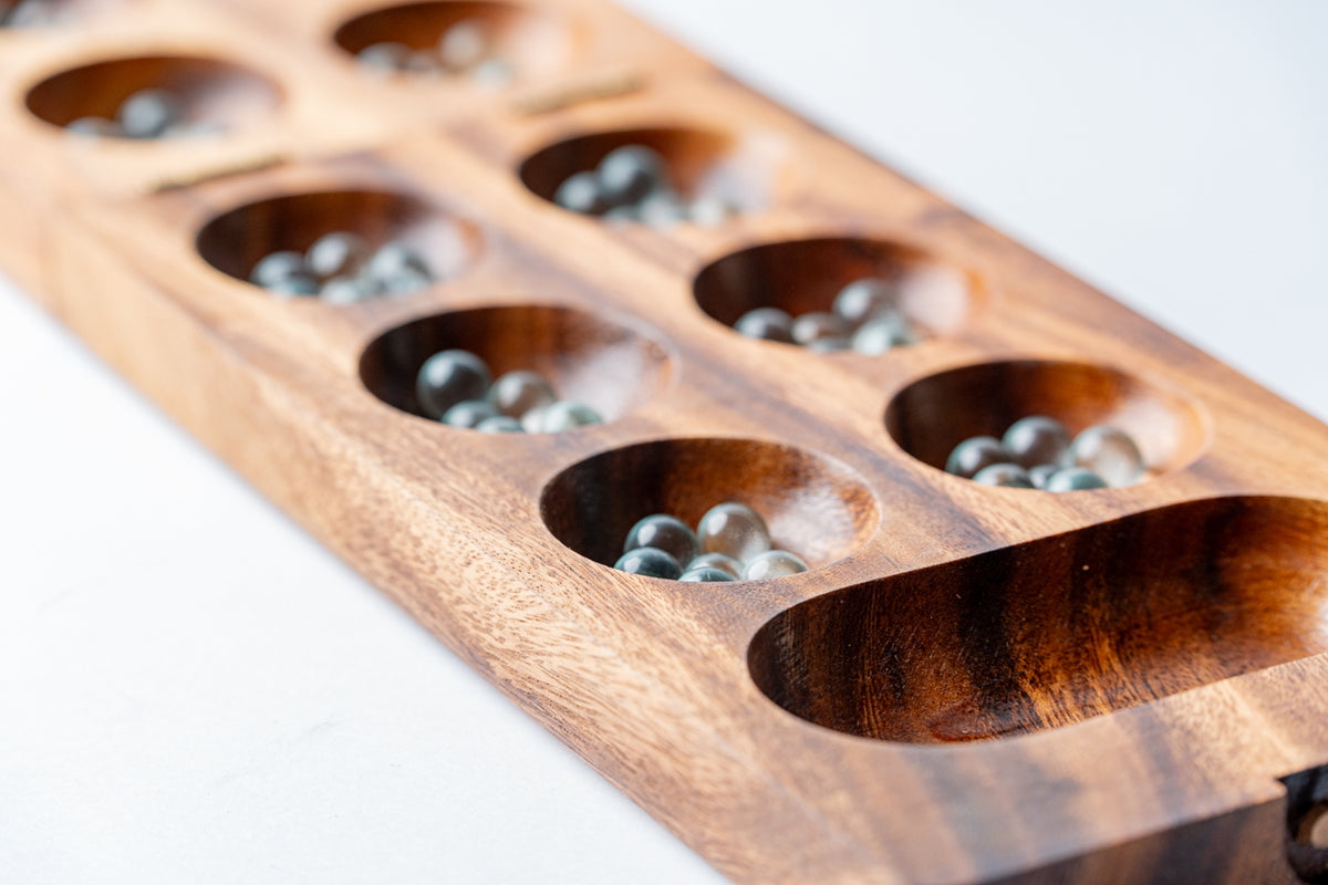 Wooden mancala board with stone playing pieces in carved cups and end storage pockets.