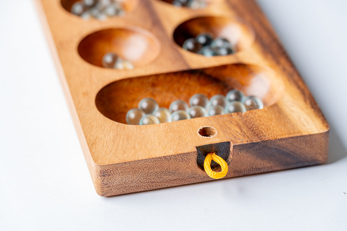 Wooden mancala board with metal game pieces in carved pockets and yellow decorative detail on edge.
