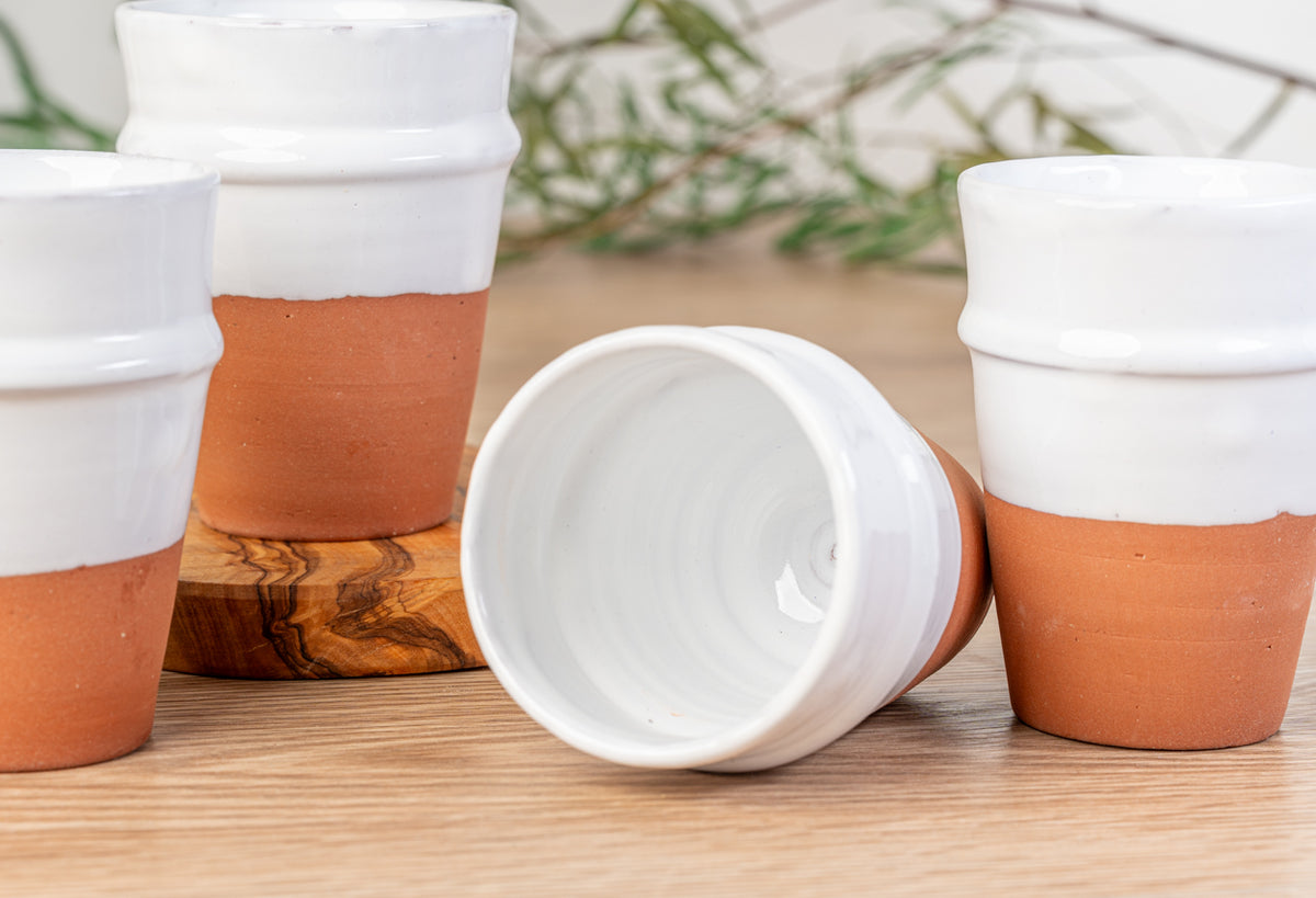 Four white ceramic cups with terracotta-colored bases displayed on wooden surface with greenery in background.