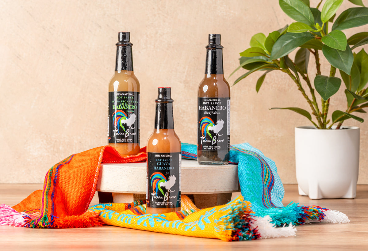 Three habanero hot sauce bottles with colorful servilleta cloth and potted plant on wooden surface.