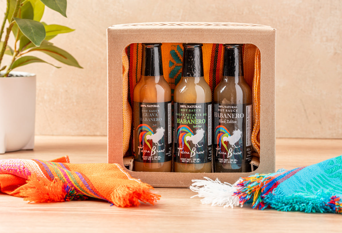 Three habanero hot sauce bottles in cardboard box with colorful striped servilleta napkins on wooden surface.