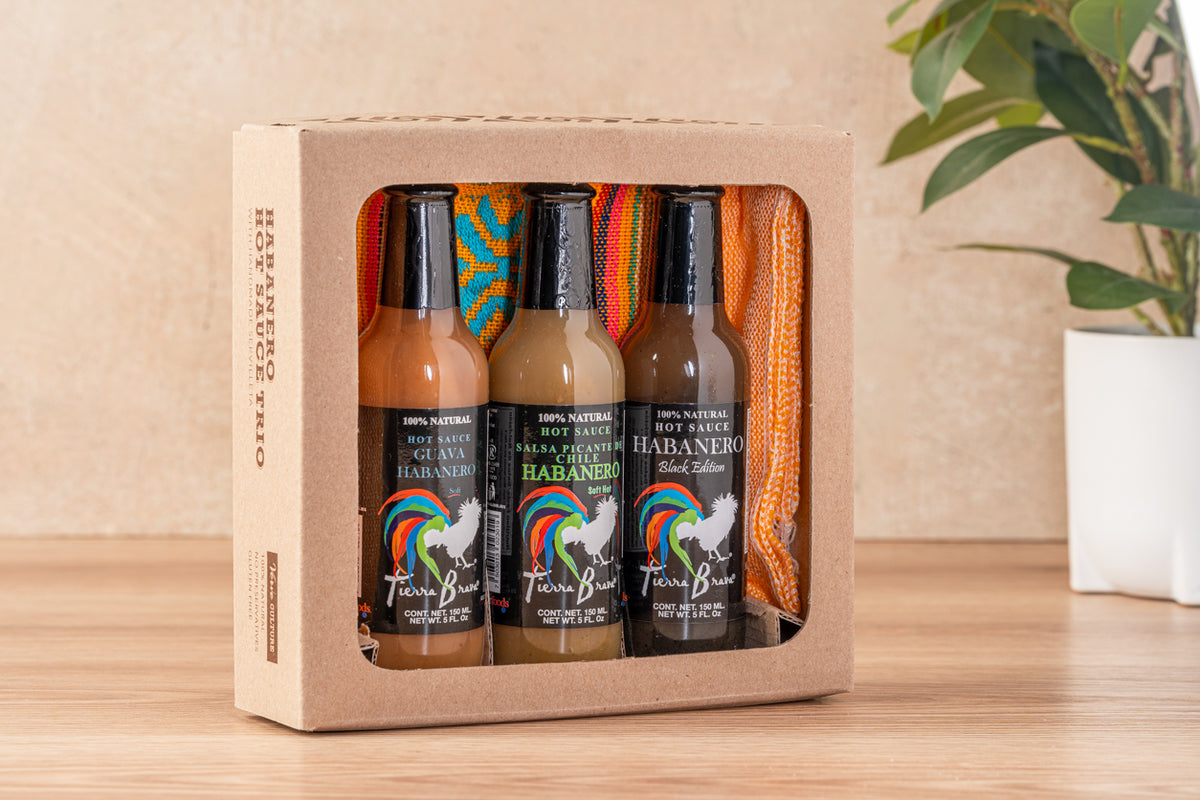 Three habanero hot sauce bottles in varying heat levels with a colorful woven servilleta, displayed in cardboard gift box.