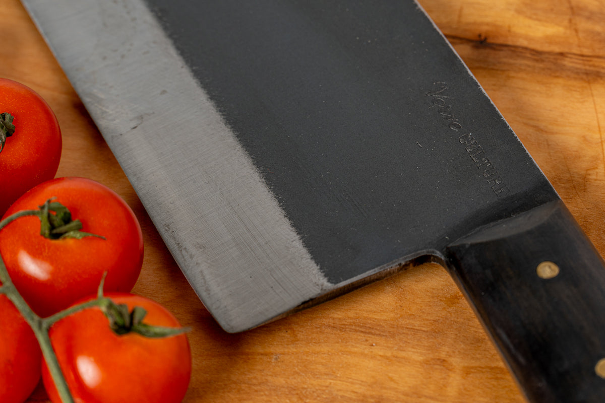 Thai cleaver knife with dark metal blade and black handle on wooden cutting board next to fresh red tomatoes.