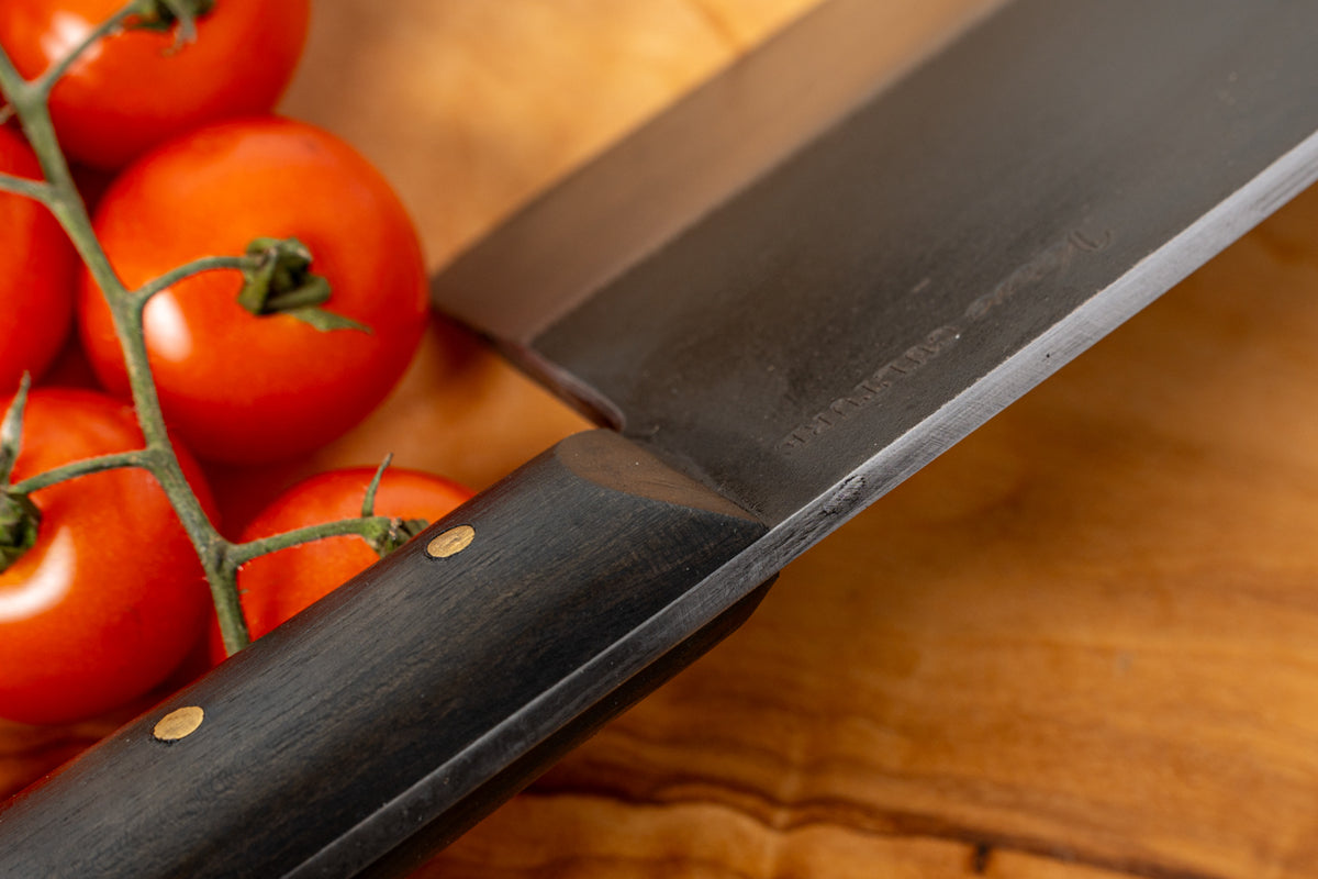 Chef's knife with dark wooden handle and metal blade next to fresh red tomatoes on vine on wooden surface.