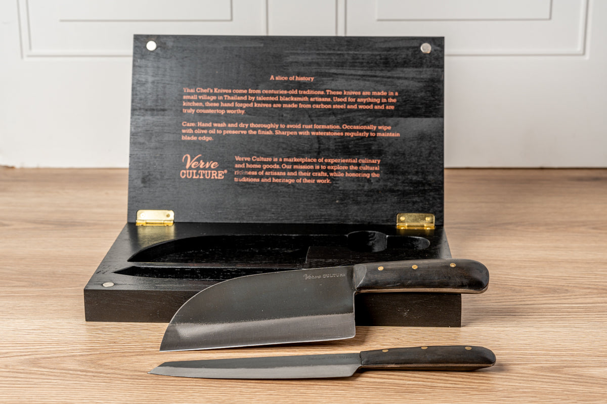 Black presentation box with two Thai knives featuring dark handles and metal blades, displayed open on wooden surface.