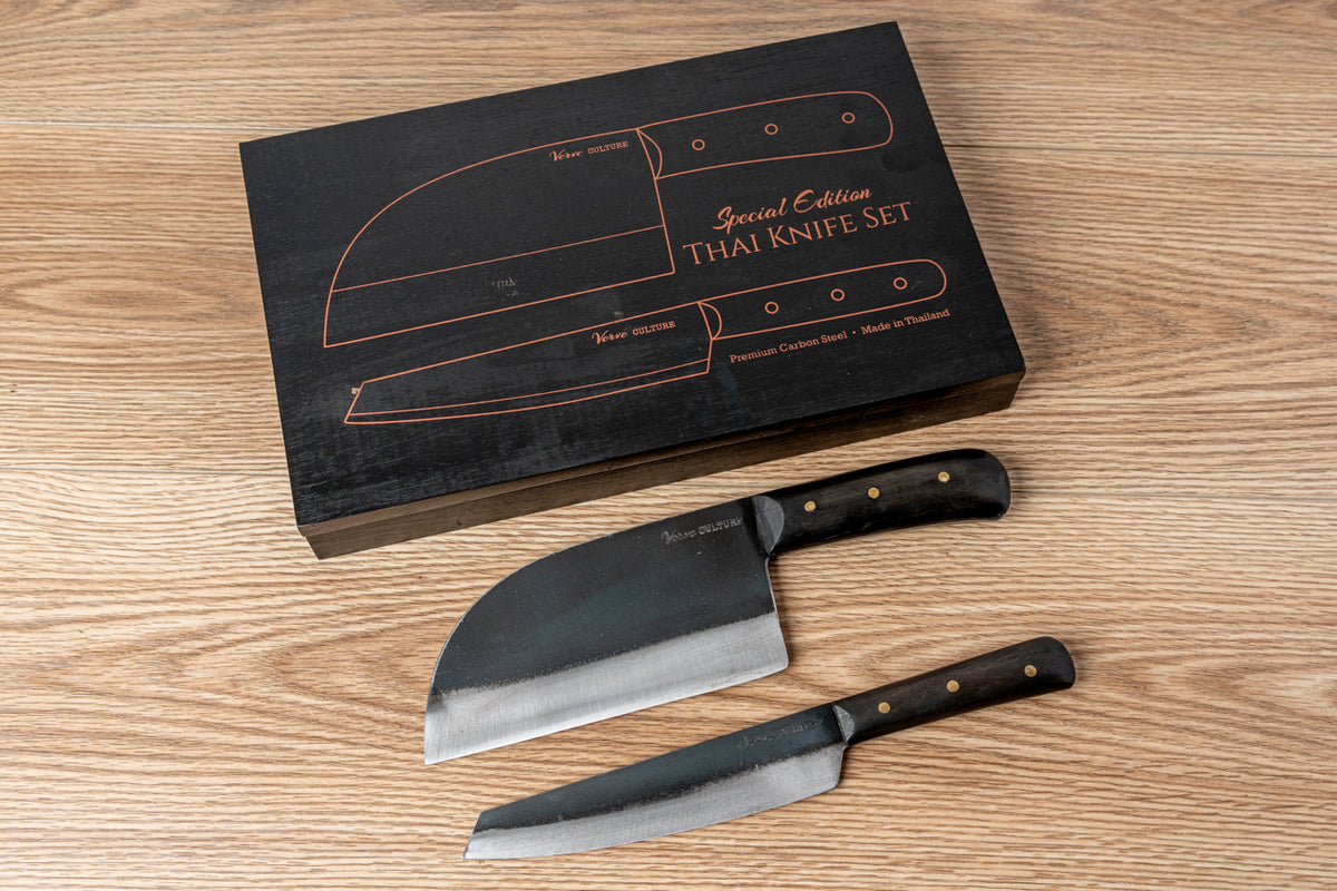 Black box with copper knife outlines containing two Thai knives with black handles and metal blades on wood surface.