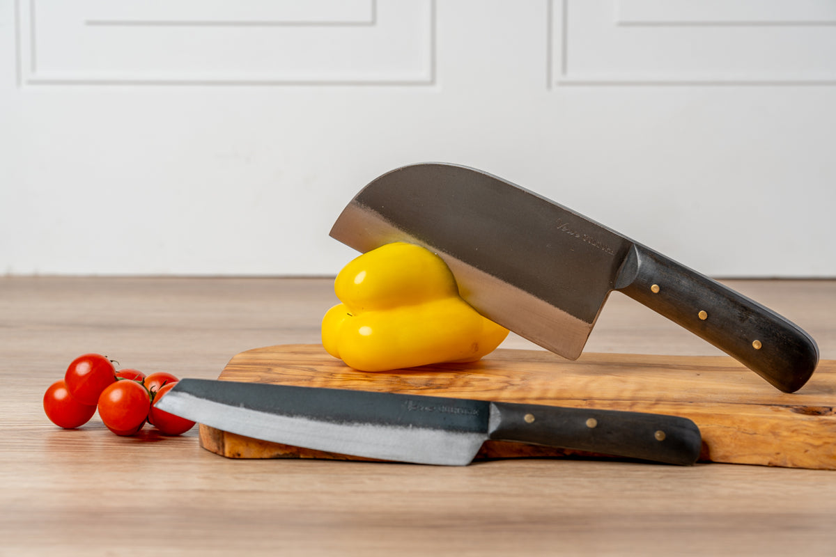 Two Thai knives with dark handles and brass rivets on a wooden cutting board, with yellow peppers and red tomatoes.