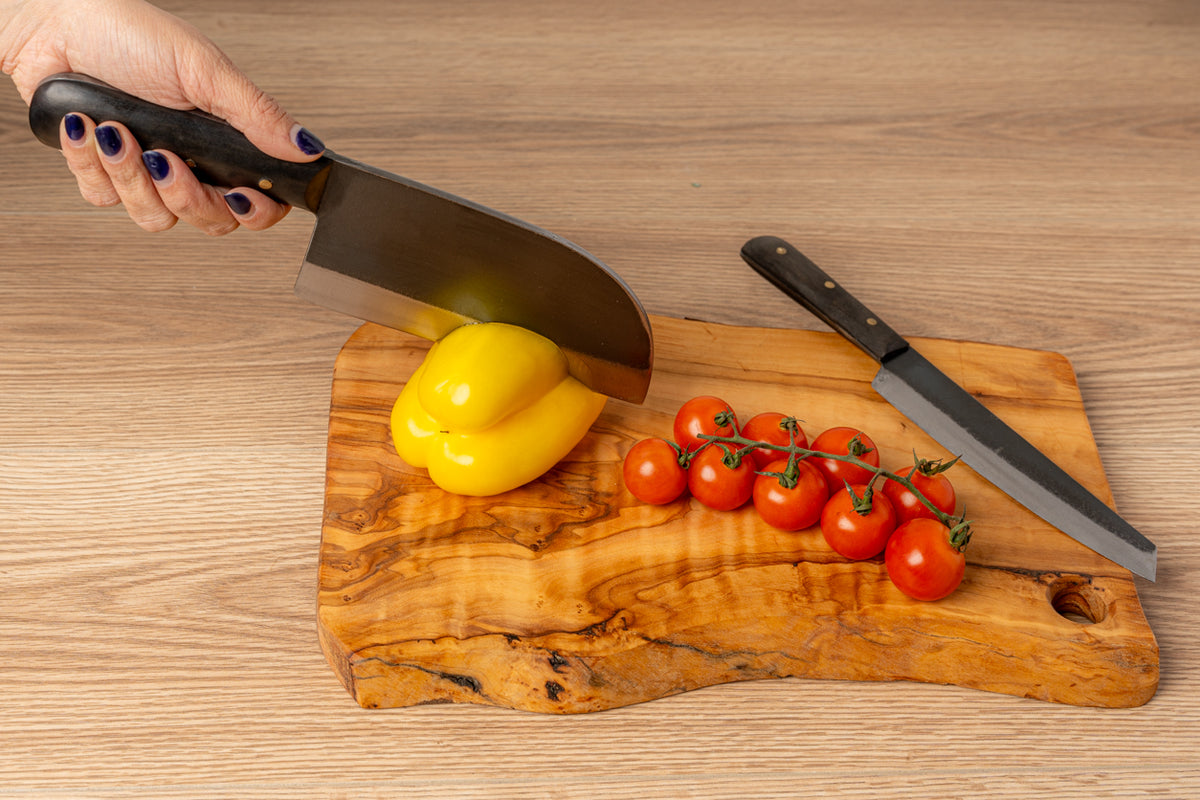 Hand holding curved black knife over wooden cutting board with yellow bell pepper, red tomatoes, and second knife.