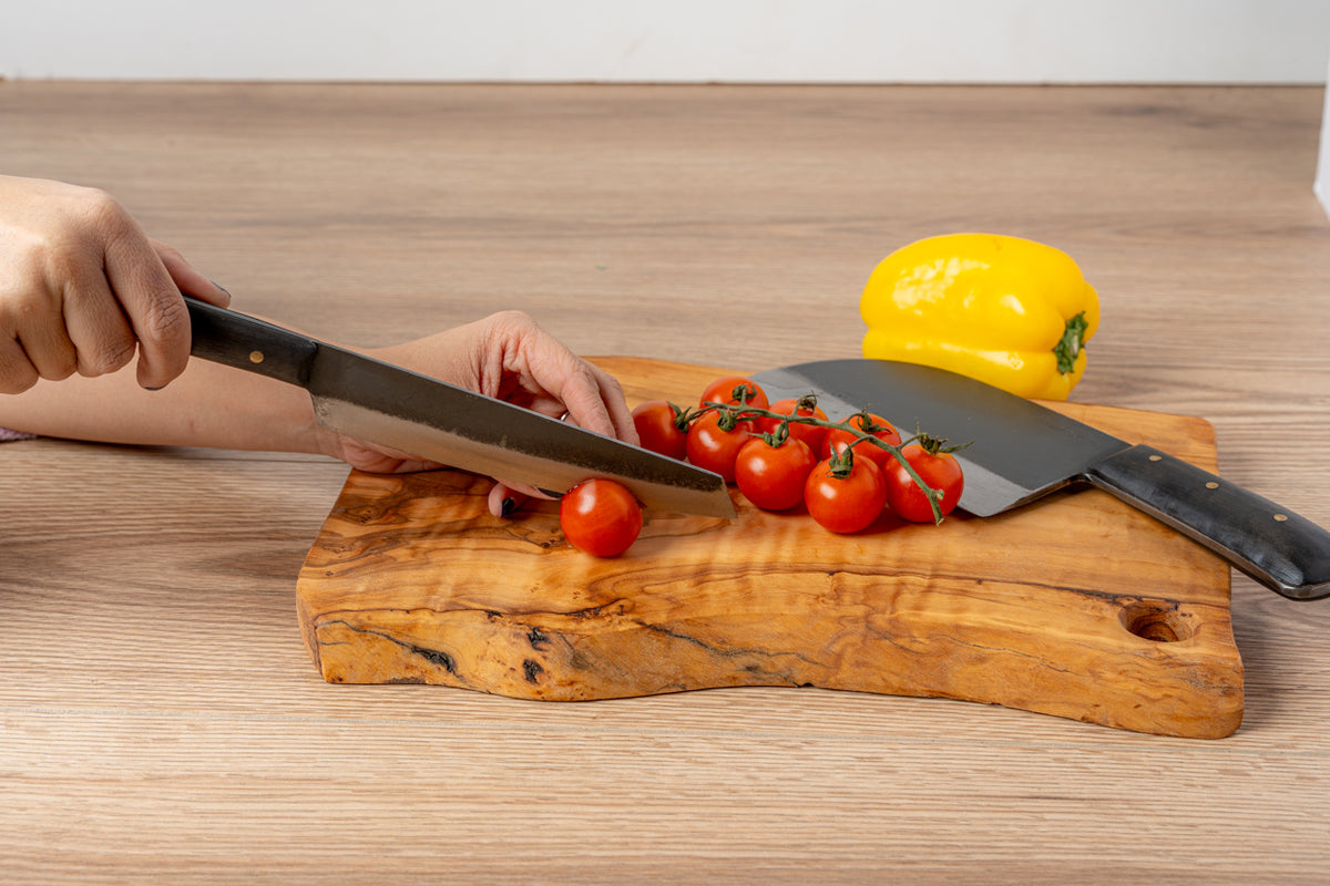 Hand holding Thai knife with black blade cutting tomatoes on wooden cutting board, yellow pepper in background.