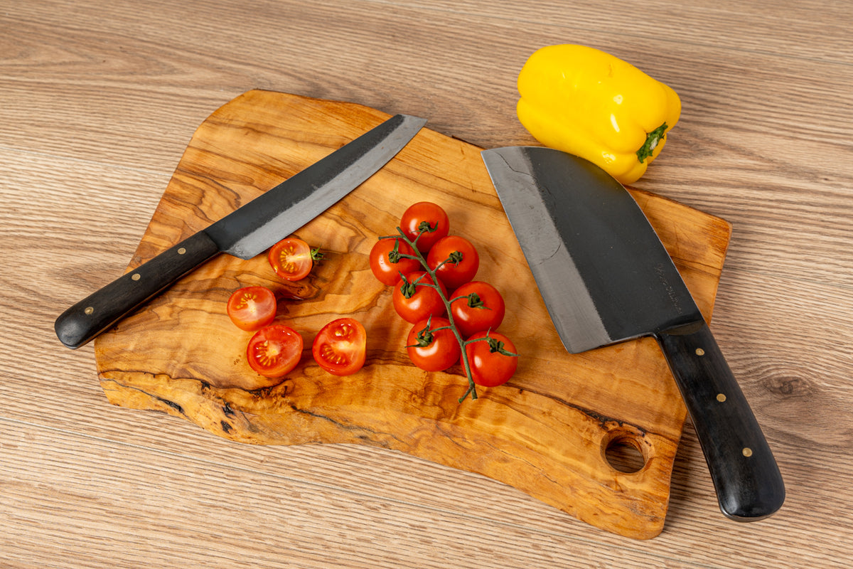 Two knives with black handles on wooden cutting board with sliced tomatoes and yellow bell pepper.