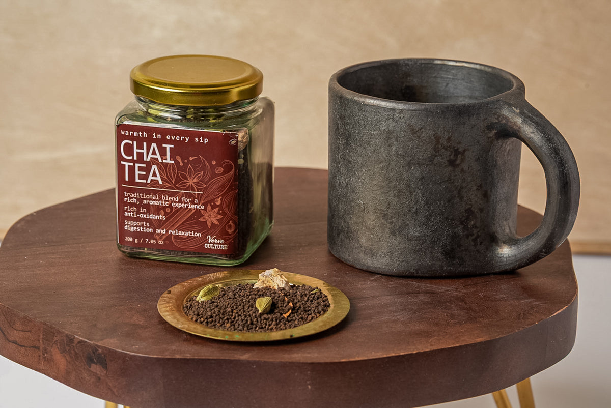 Glass jar of chai tea spice blend with gold lid, displayed with dark ceramic mug and brass bowl of whole spices on wooden tab