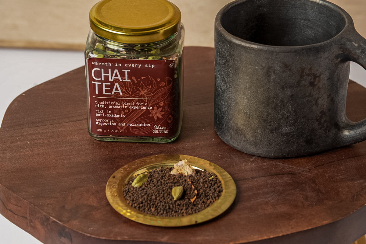 Glass jar of chai tea spice blend with gold lid next to a dark ceramic mug on a wooden surface.
